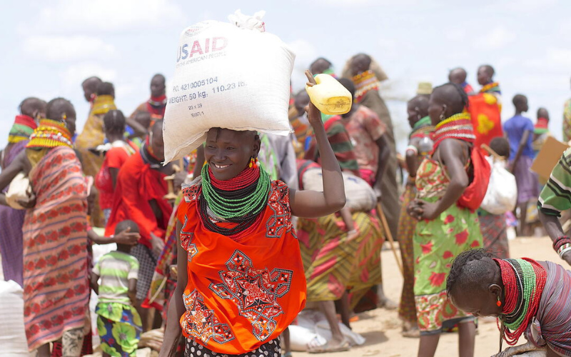 A smiling Kenyan woman carries a bag of food on top of her head, with other people in the background.