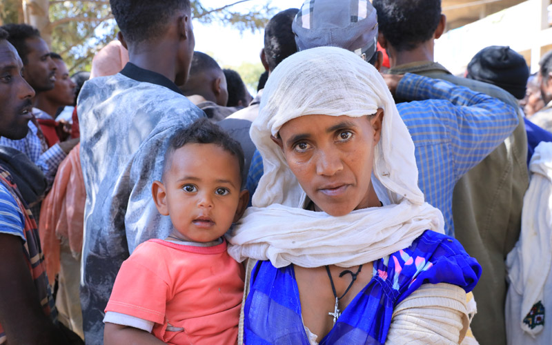 An Ethiopian woman holds a baby. There is a crowd of men and boys behind her.