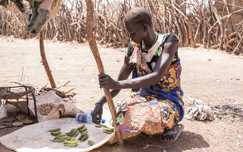 A South Sudanese mother lays out her crops on a mat under the shade of a lean-to.