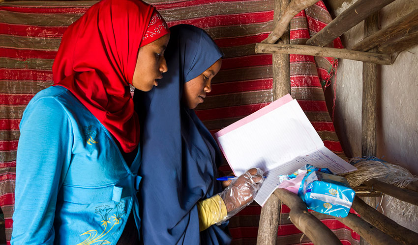 Two young women in headscarves, one writes in a notebook with an open pack of sanitary pads next to her.