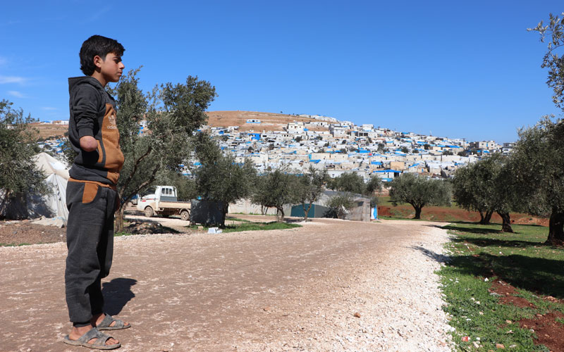 a Syrian boy stands on a dirt road. Behind him tents and makeshift structures crowd the hillside.