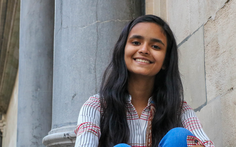 A young Bangladeshi girl smiles. She has long dark hair, and wears a plaid shirt and blue jeans.