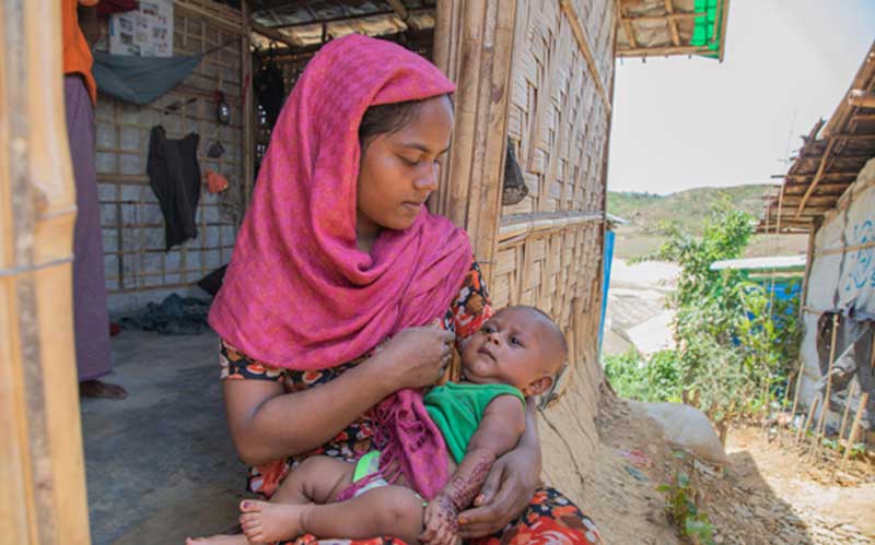 In Bangladesh’s Cox’s Bazar refugee camp, a young mother sits on the step of her hut, preparing to breastfeed her baby.