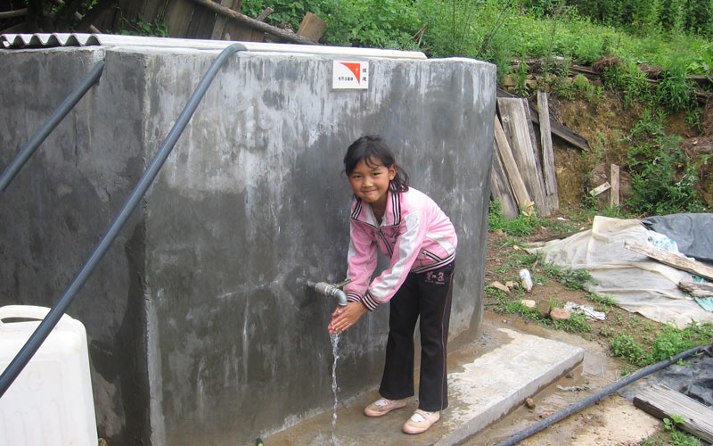 A young girl wearing a pink jacket washes her hands at a water faucet.