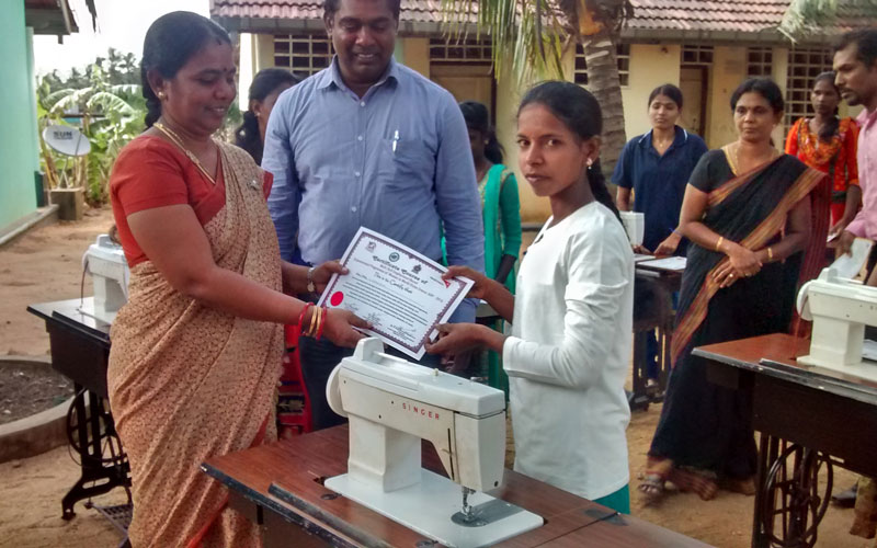 A young woman is presented with a certificate as she stands behind a white sewing machine.