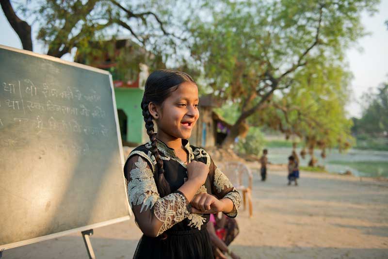 An adolescent girl in India smiles broadly while leading a class of younger students. She is outside in a rural setting, with a chalkboard behind her.