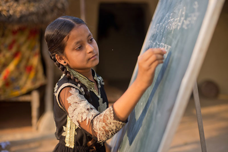 An adolescent girl in India gazes intently at the chalkboard on which she is writing. The board is propped up outside, in a rural setting.