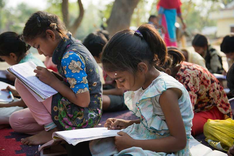 A group of young children bend intently over their writing. They are sitting on a blanket outdoors in rural India.