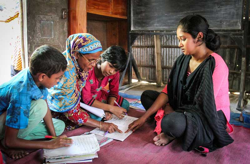 In Bangladesh, a mother and her two children hunch over a writing assignment, while a teacher points to something on the page.