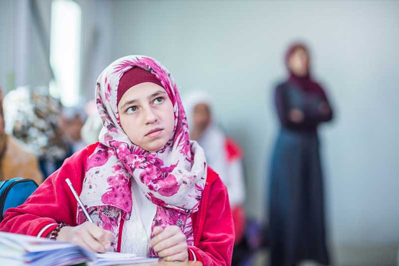 An adolescent girl in a headscarf concentrates on what the teacher is saying. She has a pencil in her right hand and a notebook in front of her.