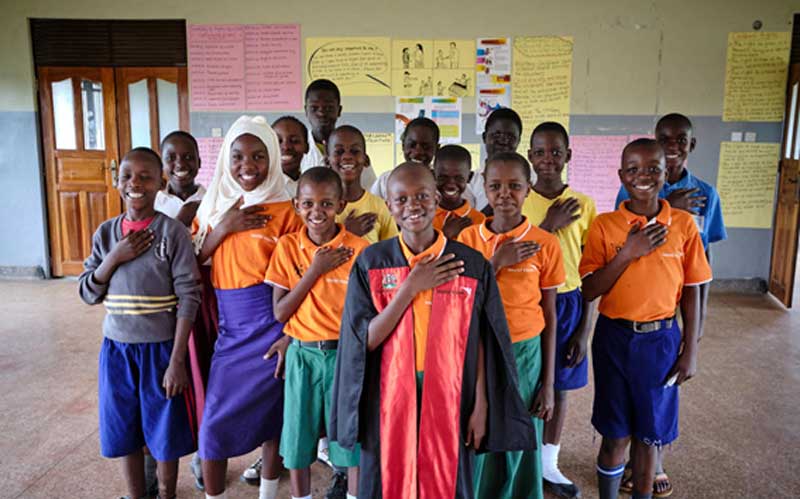 In Uganda, a group of smiling school children stand at attention for the camera, each with one hand over their hearts.