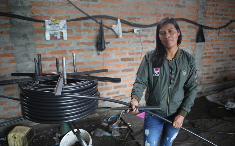 A young Ecuadorian woman smiles, holding a black hose in her hand. She's wearing jeans and a green jacket.