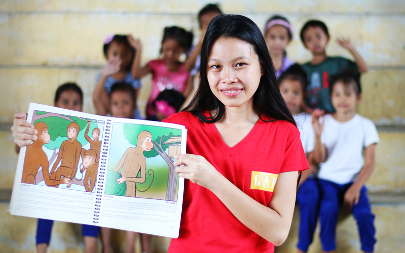 a young Philippino woman smiles. She holds a picture book and wears a red t-shirt. There are a whole group of small children seated behind her.