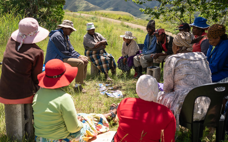 A group gathers in Lesotho. They sit outside on chairs and everyone is wearing a hat.