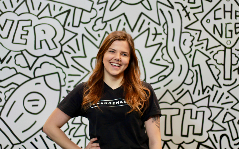 A young woman in an black tshirt that says Changemaker stands in front of a geometric print wall