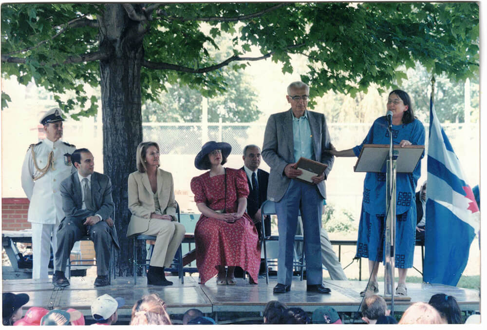 Arie Kamp is standing in the center of a stage, holding a certificate in his hands. Standing on his right, a woman in a blue dress is talking into a microphone.”
