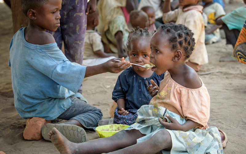 A boy kneels, to feed his sister some porridge with a spoon. They are siting on the ground.