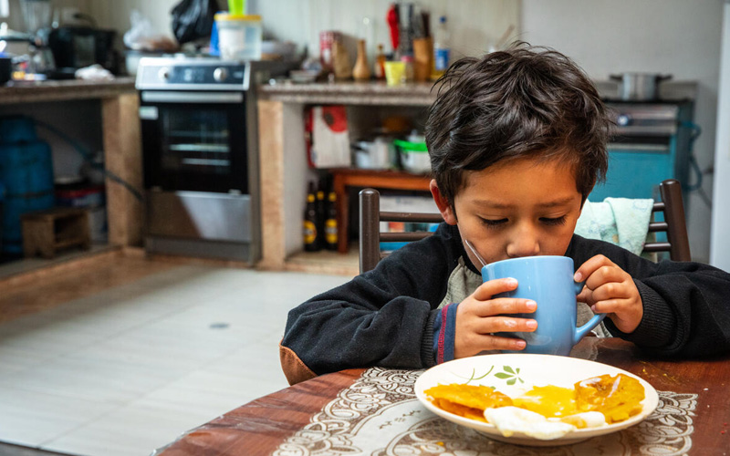 A boy sits at the kitchen table drinking from a mug.