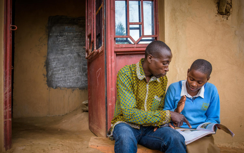 A man reads a book with his daughter, both sitting in a doorway.