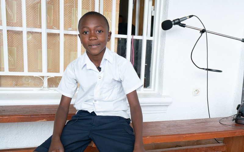 a young boy from DRC sits on bench with a microphone beside him.