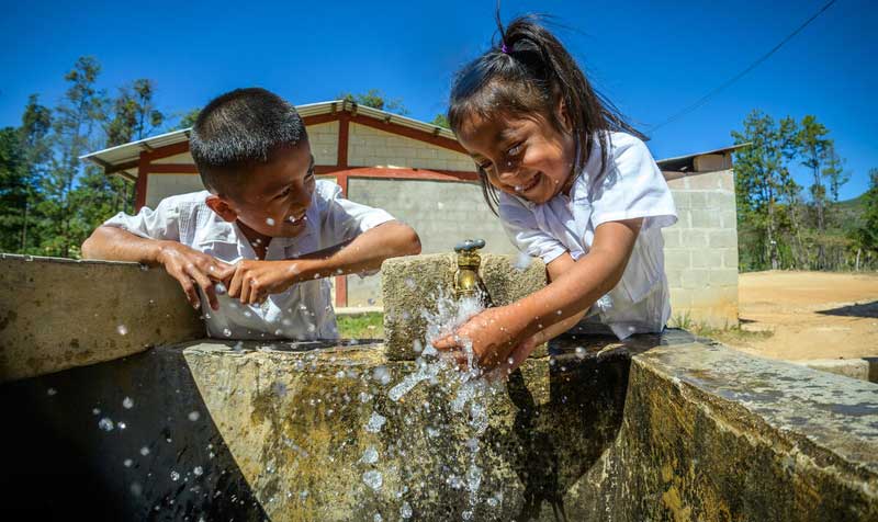 Two young children smile while washing their hands with clean water coming from an outdoor tap.