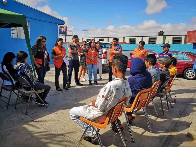 Adolescent children sit in orange chairs, facing away from the camera. One of them has a rip in his shirt. They children face a row of World Vision staff, who are talking to them.