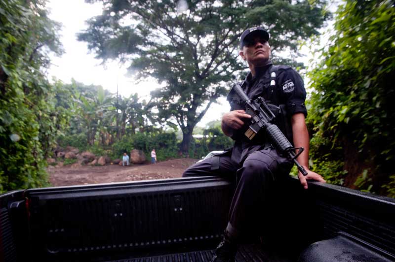 A police officer in dark uniform rides in the back of a black pick-up truck. He holds a large gun in one hand.