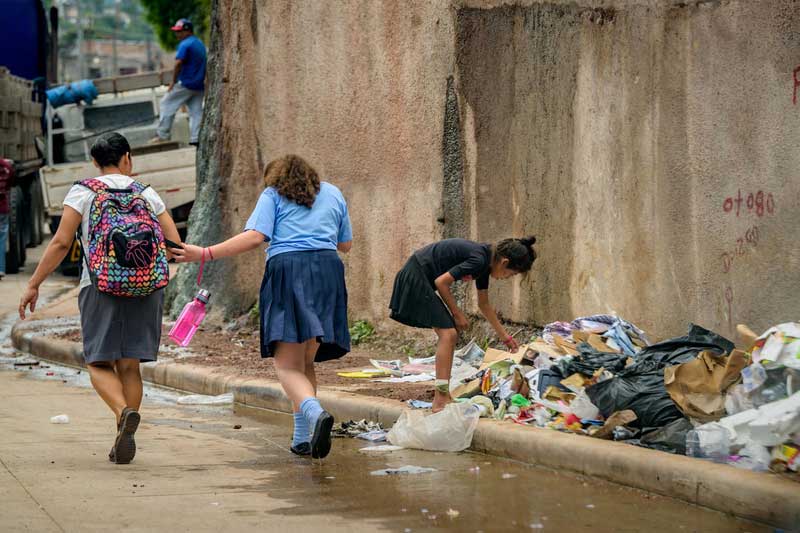 Two females walk away from the camera, heads down. To their right, a girl bends to pick through garbage at the side of the road. Behind her is a wall with graffiti.