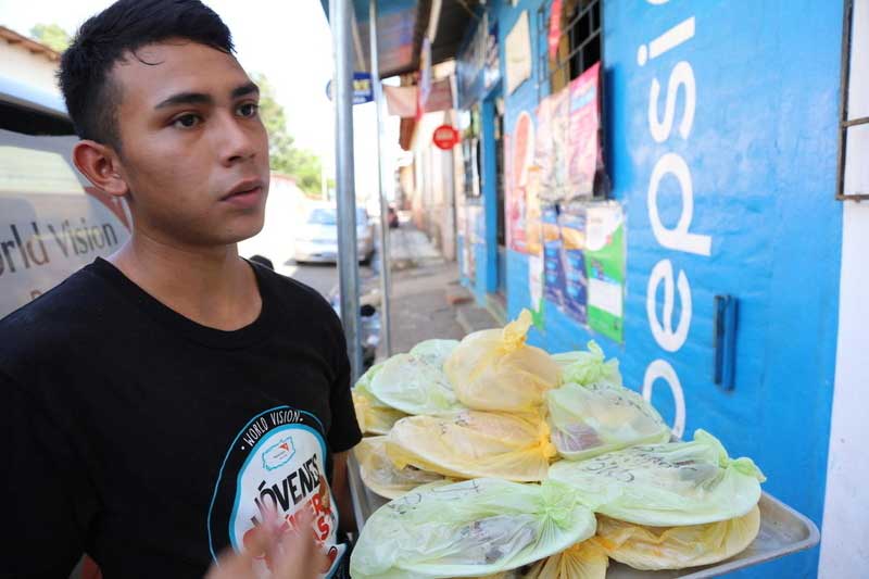 A teen or young adult boy talks to a customer. He has a large tray of wrapped tortillas on one arm.