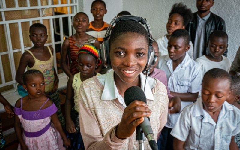 A young girl holds a microphone and wears headphones, while a group of children stand singing behind her.