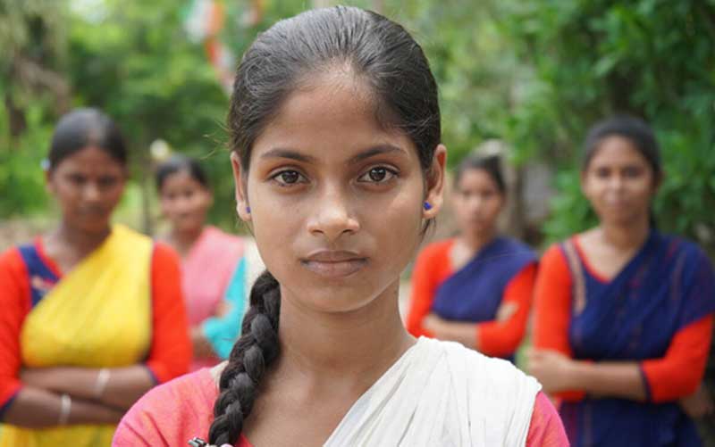 In India, a member of a girls’ advocacy group faces the camera, looking determined. Behind her stand other group members, arms folded.