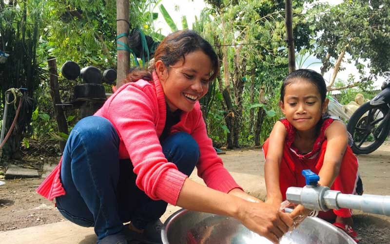 A woman and girl washing their hands at a faucet on the ground.