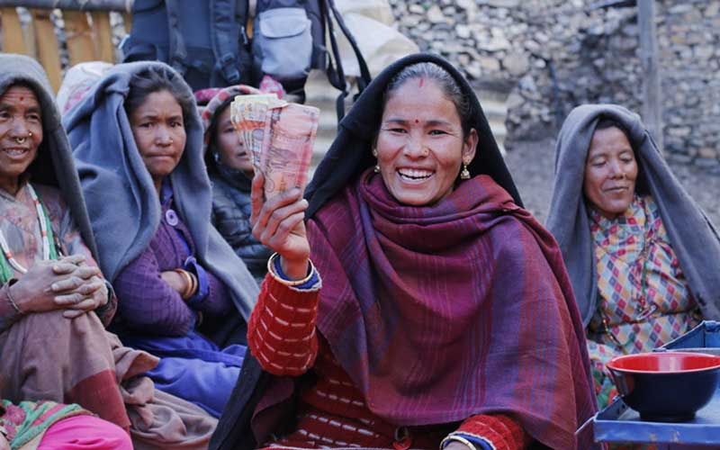 Nepal, a woman smiles proudly while holding up a handful of bills in the local currency. A group of women sit behind her.