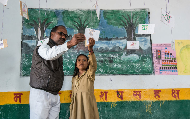 A young Indian girl stands next to her teacher in the classroom as they work on a lesson.
