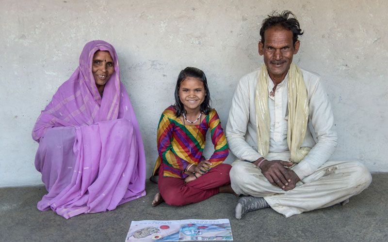 A young Indian girl sits on the floor at her home. Her parents are sitting on either side of her and they are all smiling. There is a book in front of them.