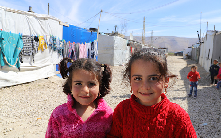 Two Syrian girls wearing bright-coloured sweaters stand side by side, smiling and looking at the camera.