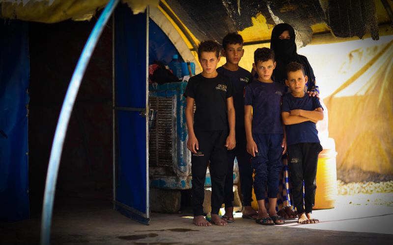 Four young Arab boys stand outside a tent, with their mother standing behind them.