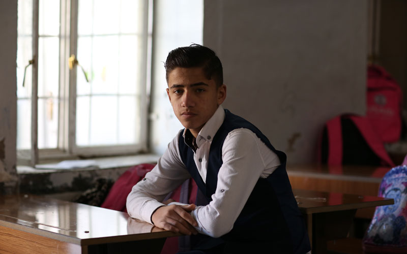 a young Iraqi boy sits at a desk and looks directly at the camera