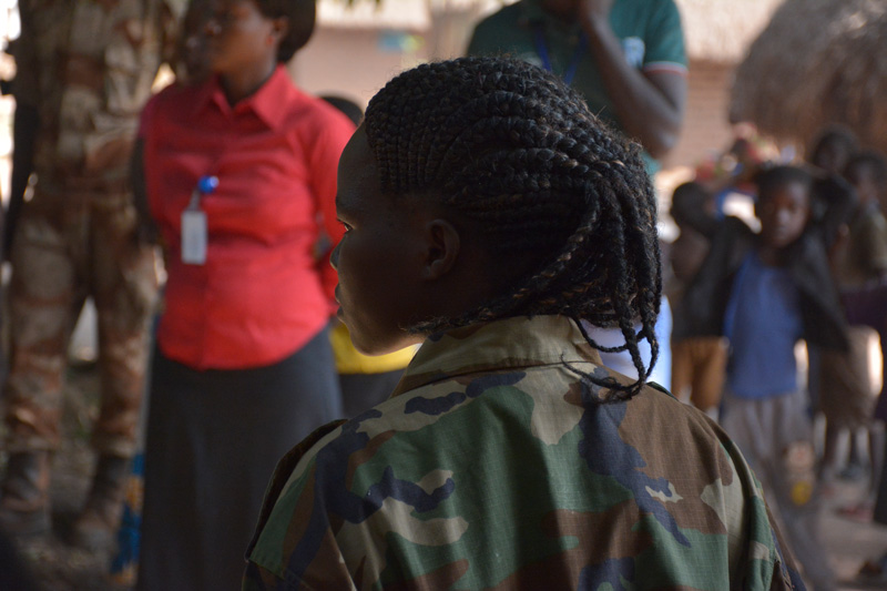 A young girl wearing military uniform stands with her back to the camera while staring to her left.