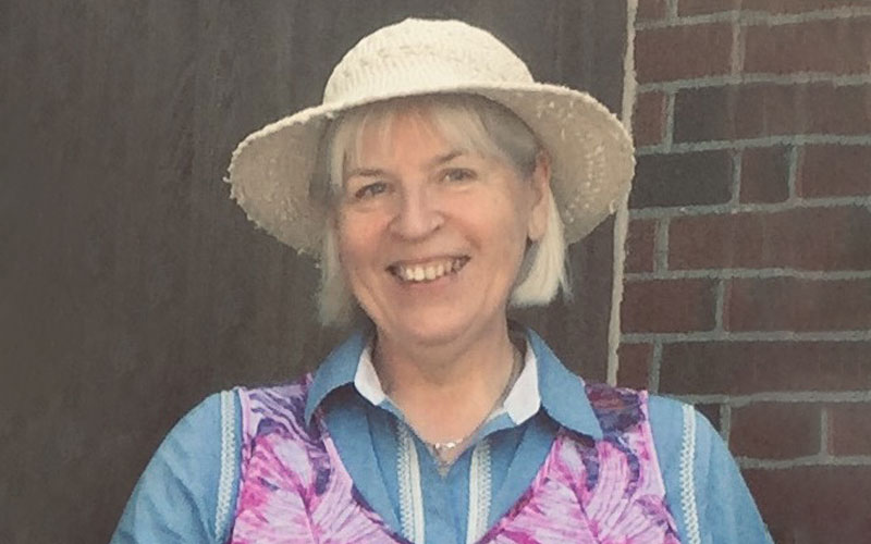 A woman standing in front of a brick wall, smiles while wearing a wide-brim sun hat.