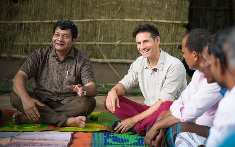 A group of men sitting on mats, talking and smiling.
