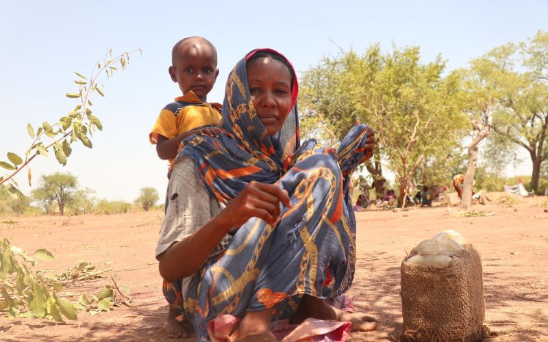 A Sudanese woman sits outside on the ground, with her child standing behind her.