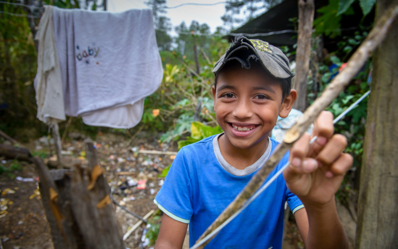 A smiling boy from Guatemala in a cap and blue shirt outside his home.