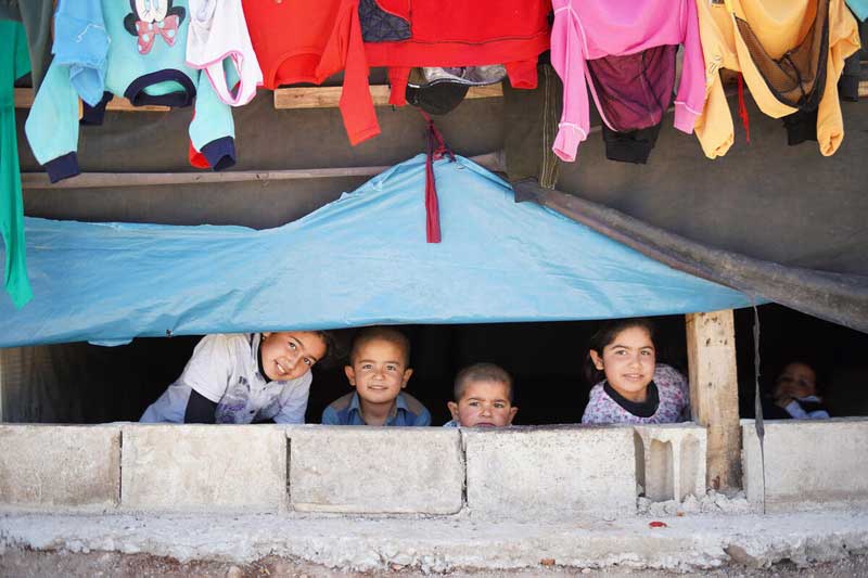 Five Syrian children under a tent with clothes hanging above them.