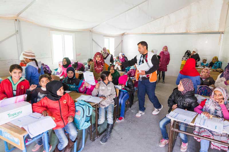 Syrian refugee children sit at school desks in a classroom set up in a refugee camp tent.
