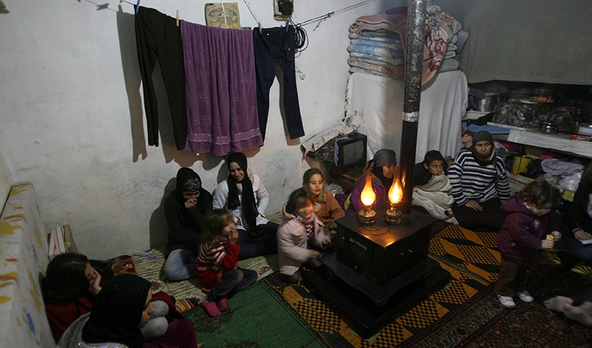 A family of Syrian refugees living in a camp, convene in a makeshift room with oil lamps and clothes hanging.