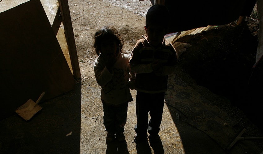 Two little children stand in a doorway in a refugee camp, and are covered by a shadow.