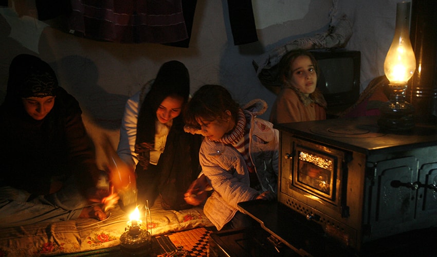 In a crowded room with an oil lamp a family plays a game together. They are Syrians living in a refugee camp.
