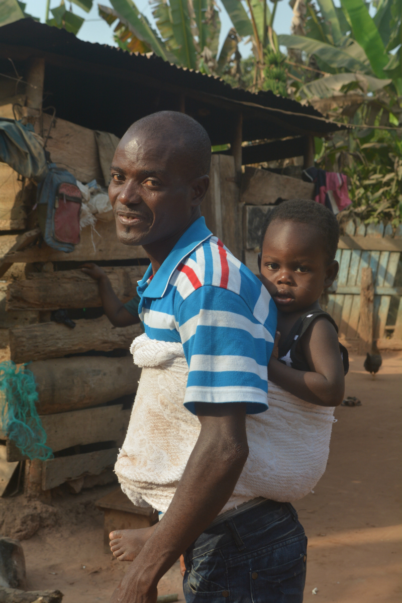 A man smiles with a baby tied to his back with cloth