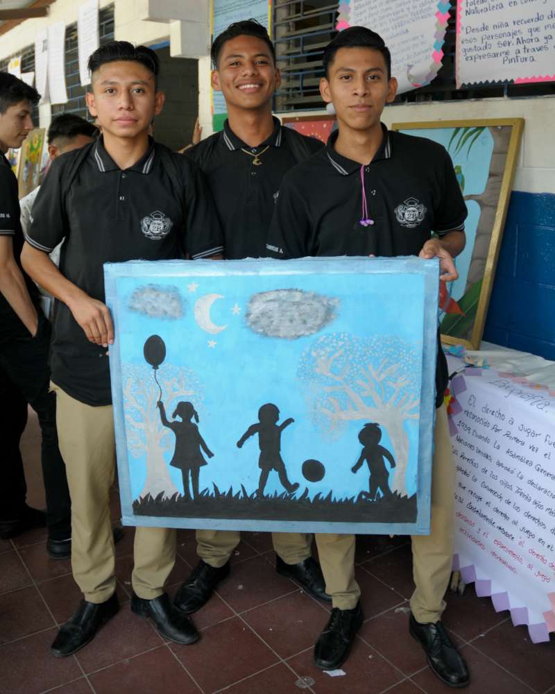 Three young men hold a painting of children playing.